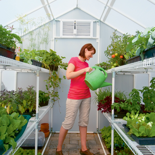 A smiling woman watering plants inside 8' X 24' solexx garden master small greenhouse
