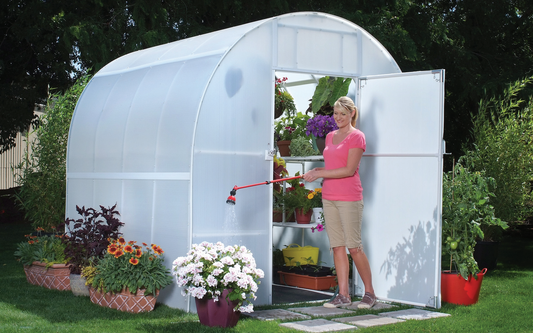 Gardener's Oasis greenhouse with a smiling woman in front of the greenhouse watering the flowers