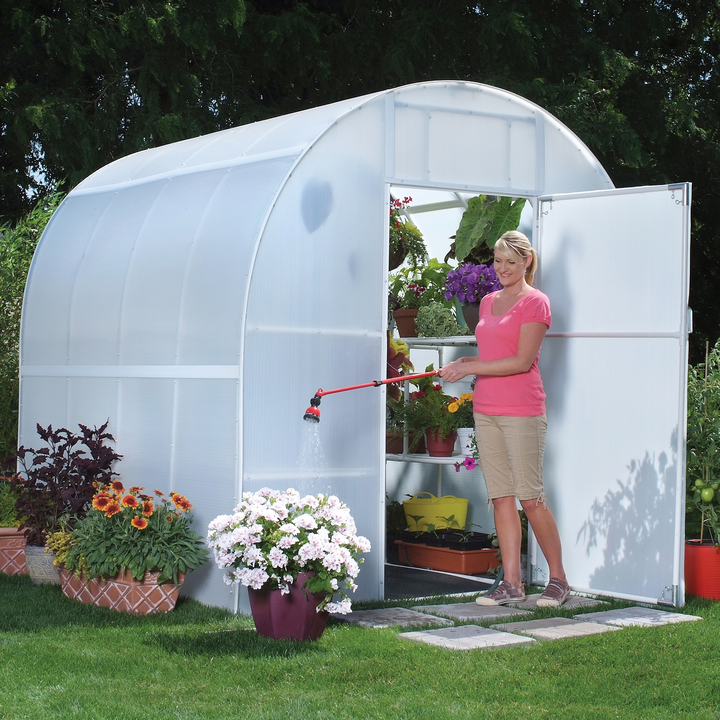 A woman watering daisy flower in front on 8' X 8' Gardener's Oasis Home Outdoor greenhouse