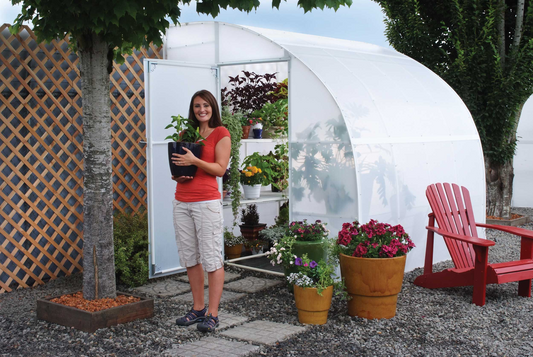 A smiling woman holding a black vase of green plant standing in front of a Wall-mounted Solexx harvester small greenhouse 