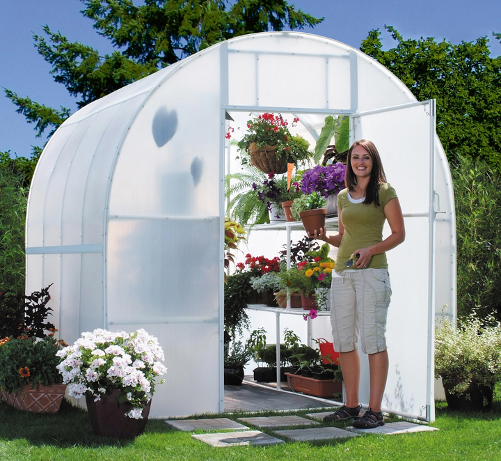 A woman smiling and holding a small brown flower vase standing in front of 8' X 12' Solexx gardeners oasis outdoor greenhouse showing flowers inside the greenhouse