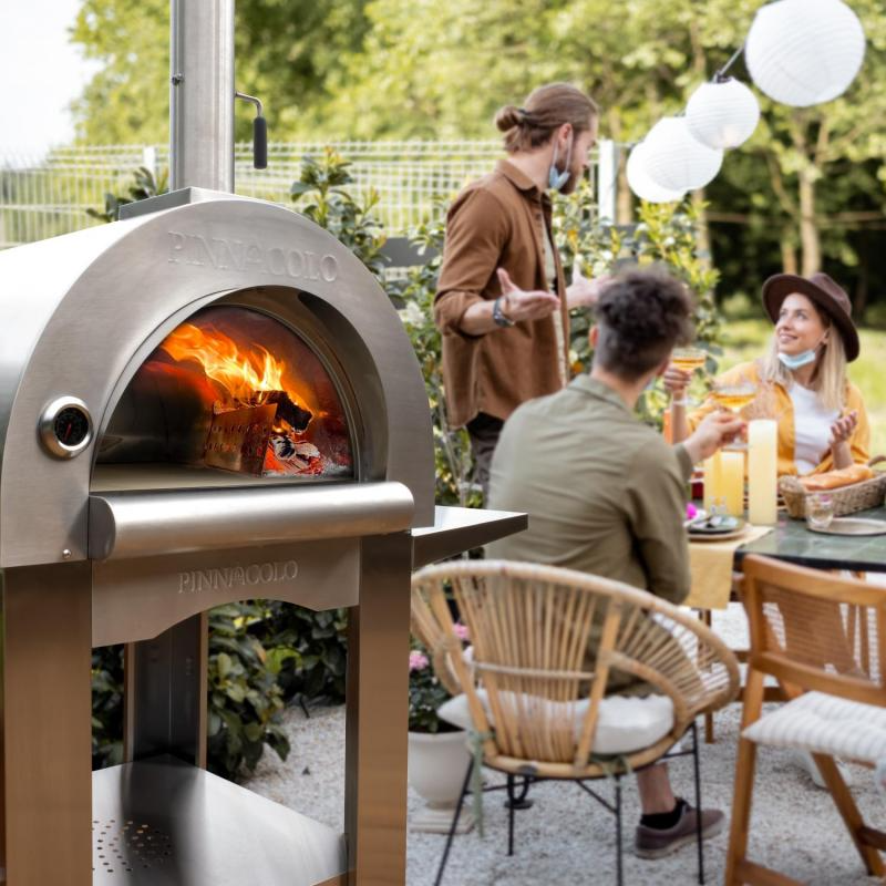 People enjoying a meal around Pinnacolo premio outdoor pizza oven in a garden setting.
